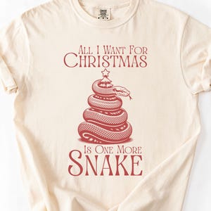 Op de afbeelding: Crèmekleurig T-shirt met een rode afbeelding. De afbeelding toont een opgerolde slang in de vorm van een kerstboom, bekroond met een ster, en de tekst "ALL I WANT FOR CHRISTMAS IS ONE MORE SNAKE."