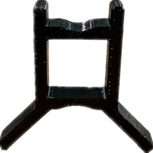 May include: A black plastic stand with a square opening in the center and three legs. The stand is designed to hold a small object.