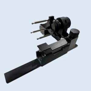 Puede incluir: Un dispositivo de metal negro con un brazo largo y una cabeza giratoria con tres puntas. El dispositivo parece ser una herramienta o instrumento de precisión.