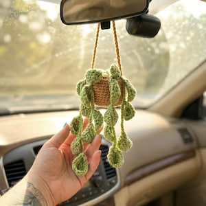 May include: A crocheted hanging plant ornament featuring a small tan pot and multiple trailing stems with light green leaves. The plant is suspended by a braided tan yarn cord, designed to hang from a rearview mirror in a vehicle interior.