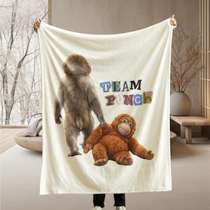 May include: Cream-colored blanket featuring two cartoon orangutans and the text "TEAM PUNCH" in colorful letters. One orangutan is standing, and the other is lying down. The blanket is held up by a person.