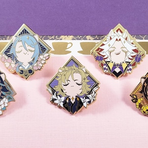 May include: Five diamond-shaped enamel pins featuring anime-style character portraits. Each pin has a gold border and a unique color scheme, including blue, white, purple, and yellow. The pins are set against a pink surface.