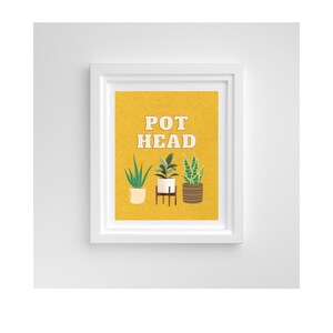 Puede incluir: Una impresión amarilla con el texto "POT HEAD" en letras blancas. Se muestran tres plantas en macetas debajo del texto. Las plantas están en macetas blancas, marrones y blancas. La impresión está enmarcada en un marco blanco.