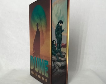 May include: A hardcover book with a gold and brown cover featuring a colorful illustration of a desert landscape with two figures. The title "Dune" and the author "Frank Herbert" are printed on the cover.