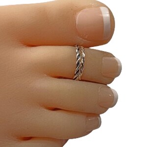 May include: A silver-colored toe ring with a braided design. The ring is worn on a foot with a light skin tone, and the toenails are painted with a French manicure.