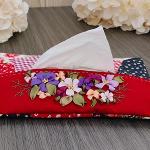 May include: A decorative tissue box cover with a red front panel, embellished with embroidered flowers in purple, white, and peach. The sides display patchwork patterns in red, white, and blue. White tissues are visible.