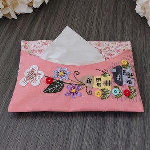 Puede incluir: Funda de caja de pañuelos de tela rosa con bordados florales y aplicaciones. La funda presenta flores bordadas, hojas y casitas. La abertura superior revela pañuelos blancos. El forro interior tiene un estampado floral rosa.