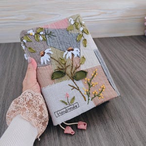 Puede incluir: Una funda de libro de patchwork hecha a mano con bordado floral. La funda presenta una mezcla de telas de tonos neutros, adornada con flores blancas parecidas a margaritas, hojas verdes y detalles amarillos. La palabra "Handmade" está bordada en la funda.