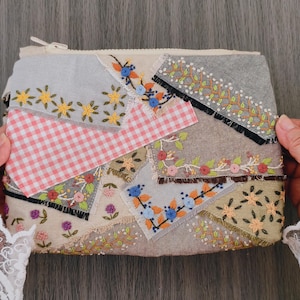 May include: A rectangular zippered pouch featuring a patchwork design of various fabric scraps. The surface includes light gray, beige, and brown fabric pieces with embroidered floral patterns in yellow, blue, pink, and green, plus a pink gingham section.