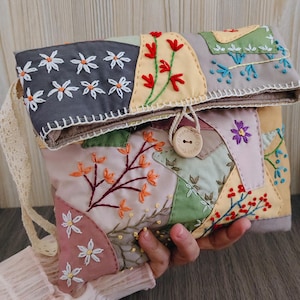 May include: A patchwork clutch with floral embroidery in various colors. The bag features a button closure and a cream-colored lace strap. The design includes embroidered flowers, leaves, and berries on a mix of pastel and neutral fabric patches.