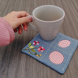 Puede incluir: Una taza de cerámica blanca descansa sobre un posavasos cuadrado de mezclilla. El posavasos presenta flores rosas y hojas verdes bordadas, con dos recortes circulares rellenos de tela de cuadros y rayas rosas y blancas.