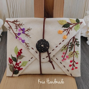 May include: Cream-coloured envelope clutch bag with embroidered floral patterns in red, purple, yellow, and green. It fastens with a brown leather cord and a dark button. The text "Knia Handmade" is visible.