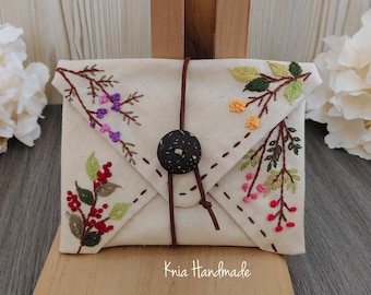 Può includere: Pochette beige a forma di busta con dettagli floreali ricamati in rosso, viola, giallo e verde. La pochette è assicurata con un cordino in pelle marrone e un bottone scuro. Il testo "Knia Handmade" è visibile.
