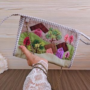 Puede incluir: Un bolso acolchado hecho a mano con una escena de paisaje con casas y árboles. El bolso tiene una combinación de colores marrón, rosa y verde, con un borde a cuadros y una correa de cadena dorada. El diseño es una aplicación bordada.
