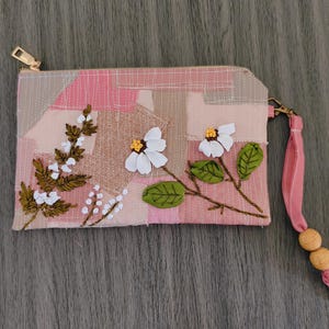 May include: A rectangular zippered pouch featuring a patchwork design in shades of pink and beige with white stitching. The front is decorated with embroidered white flowers, green leaves, and brown stems. A pink wrist strap with two wooden beads is attached.
