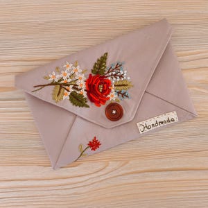 May include: A taupe envelope-style clutch with an embroidered floral design. The embroidery features a large red rose, white and blue flowers, and green leaves. A brown button secures the flap, and a small tag reads "Handmade".