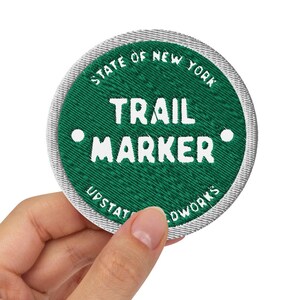 May include: A green embroidered patch with white text that reads "STATE OF NEW YORK TRAIL MARKER" with a white dot on either side of the word "TRAIL". The patch has a silver border and the text "UPSTATE OUTWORKS" is printed in white on the silver border.
