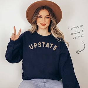 以下が含まれることがあります： 前面にタンカラーで「UPSTATE」という文字がプリントされたネイビーブルーのスウェットシャツ。ブラウンの帽子をかぶった人が着用しています。