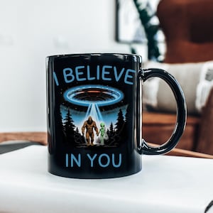 May include: Black coffee mug with a white graphic of a Bigfoot and an alien standing together under a UFO. The text "I Believe in You" is printed on the mug.