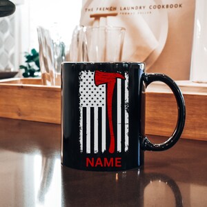 May include: Black ceramic mug with a white and red American flag design featuring a red axe. The text "NAME" is printed below the flag.