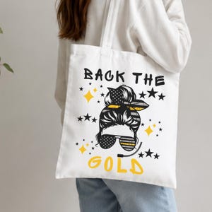 Puede incluir: Bolso tote blanco con un gráfico de una mujer con gafas de sol y una diadema, rodeado de estrellas y diamantes amarillos. El texto "BACK THE GOLD" está impreso en negro y amarillo.