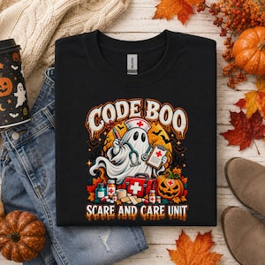 May include: A black crewneck t-shirt featuring a graphic of a ghost dressed as a nurse with a stethoscope, syringe, and clipboard. The text reads CODE BOO SCARE AND CARE UNIT, accompanied by a medical kit, medicine bottles, and a jack-o'-lantern.