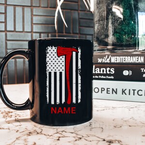 May include: Black ceramic coffee mug featuring a white and red American flag design with a red axe graphic. The text "NAME" is printed on the mug.