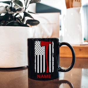 May include: Black ceramic mug featuring a white and red American flag design with a red axe graphic. The mug has white text that says "NAME" at the bottom.