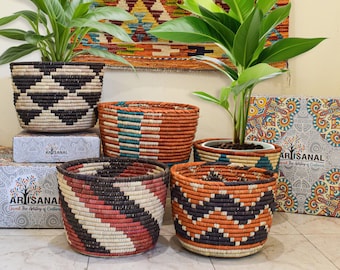 May include: Five woven baskets in various colors and patterns, including black, white, red, orange, and blue. The baskets are arranged on a tiled floor in front of a wall with a colorful rug hanging on it. The text "Artisan" is visible on the boxes behind the baskets.