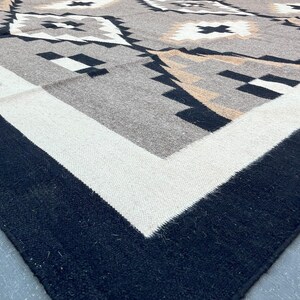 May include: A close-up of a woven rug with a geometric pattern. The rug features a border of black and white, with a central design in shades of gray, tan, and black. The pattern includes diamond and cross shapes.