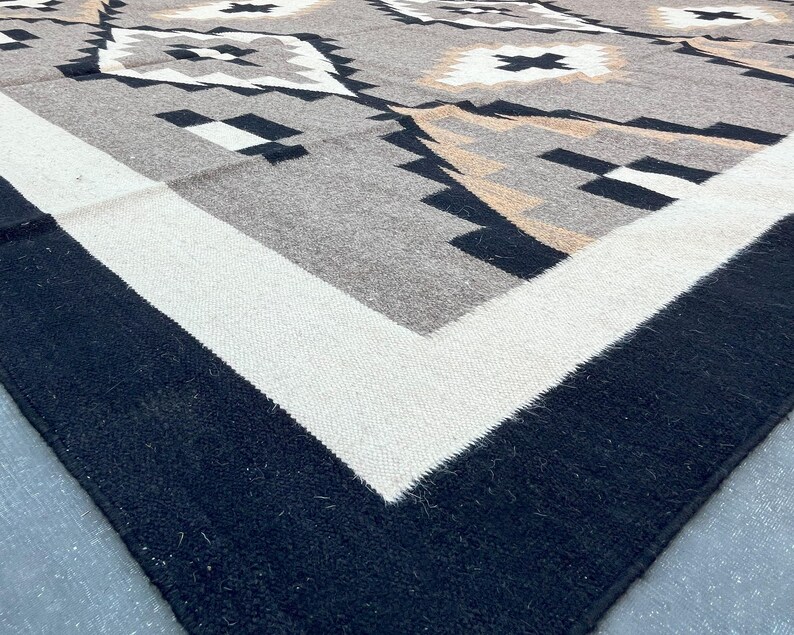 May include: A close-up of a woven rug with a geometric pattern. The rug features a border of black and white, with a central design in shades of gray, tan, and black. The pattern includes diamond and cross shapes.