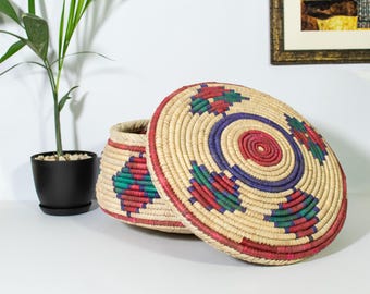 May include: A woven storage basket with a lid, featuring a colorful geometric pattern in shades of red, blue, and green. The basket is made of natural materials and has a round shape.