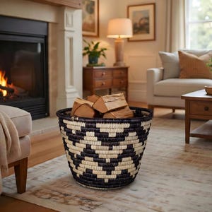 May include: A woven basket with a black and cream geometric pattern, filled with firewood, sits on a rug in front of a fireplace. The fireplace has a burning fire inside. A beige ottoman and a sofa are in the background.