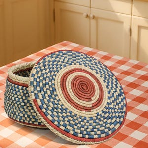 May include: A round, lidded basket crafted from coiled fibers in a woven pattern. The design features concentric circles of red, cream, and blue, with a matching blue and cream checkered pattern on the sides of the basket and the lid's edge.