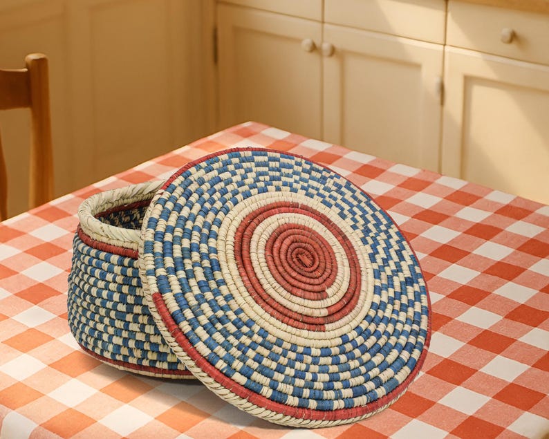 May include: A round, lidded basket crafted from coiled fibers in a woven pattern. The design features concentric circles of red, cream, and blue, with a matching blue and cream checkered pattern on the sides of the basket and the lid's edge.
