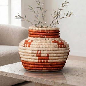 Può includere: Un vaso in vimini intrecciato con un design geometrico beige naturale e arancione. Il vaso è riempito di rami frondosi e poggia su un tavolo di pietra. Il vaso ha una forma rotonda e un'ampia apertura.