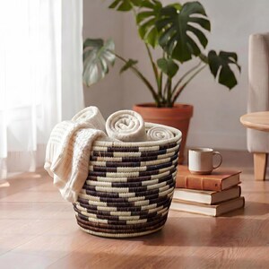 May include: A round woven basket with a geometric pattern contains rolled and folded cream blankets. The basket is on a wooden floor, near a potted plant and a small side table. A mug and books are also visible.