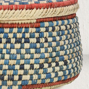 May include: A round woven basket with a lid, featuring a repeating pattern of natural beige, royal blue, and navy blue fibers. The basket includes horizontal bands of vibrant red at the top of the lid and the base, constructed from coiled material.
