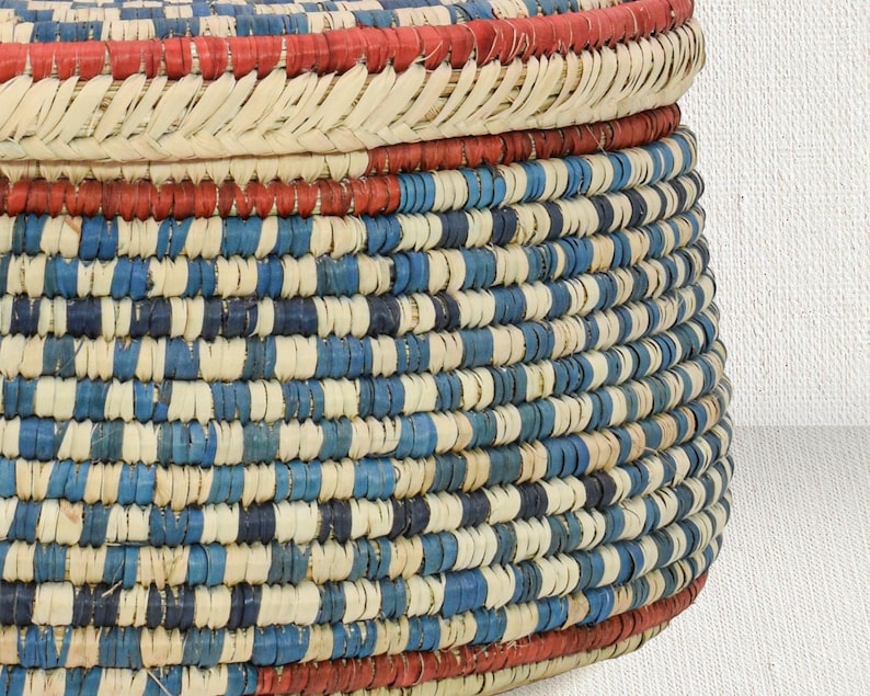 May include: A round woven basket with a lid, featuring a repeating pattern of natural beige, royal blue, and navy blue fibers. The basket includes horizontal bands of vibrant red at the top of the lid and the base, constructed from coiled material.