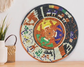 May include: A round, woven wall basket with a vibrant, geometric design. The basket features a black border and a central spiral pattern in shades of orange, yellow, green, and blue. The design includes stylized figures and shapes.