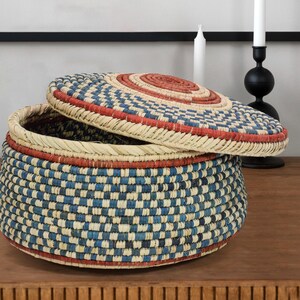 May include: A round woven basket with a matching lid, featuring a repeating checkered pattern in shades of blue, cream, and terracotta red. The basket has a coiled construction with a flat base and a slightly domed lid that rests partially open.
