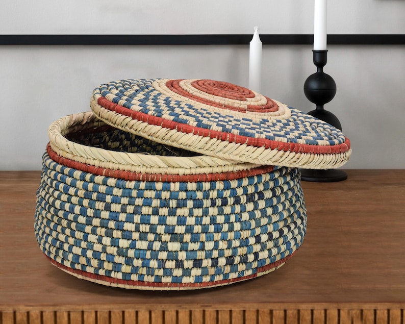 May include: A round woven basket with a matching lid, featuring a repeating checkered pattern in shades of blue, cream, and terracotta red. The basket has a coiled construction with a flat base and a slightly domed lid that rests partially open.