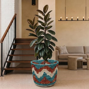 May include: A large potted plant with dark green leaves sits in a woven basket with a geometric pattern of red, white, and teal. The basket is on a light-colored floor in a modern living space. A staircase and sofa are in the background.