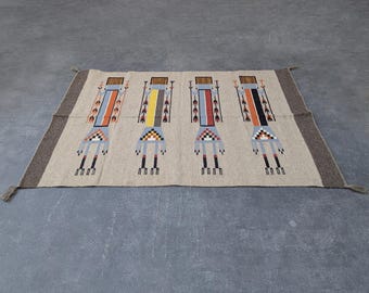 May include: Rectangular beige rug with four stylized figures in the center. Each figure has a rectangular head, a body with vertical stripes in various colors, and geometric patterns at the feet. The rug has a brown border and tassels at the ends.