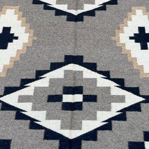 May include: A gray, white, black, and tan geometric patterned rug. The rug features a repeating diamond design with a cross in the center of each diamond. The rug is made of a soft, woven material.