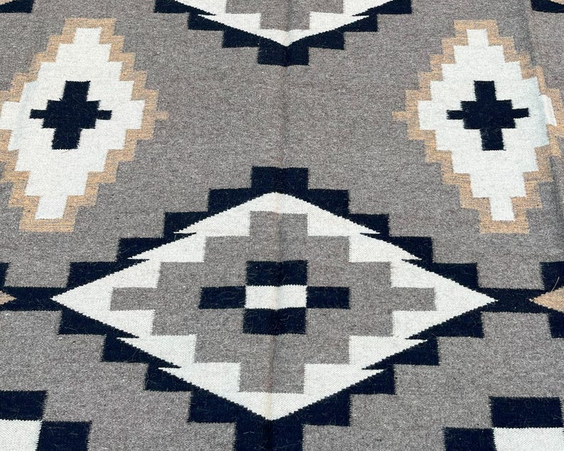May include: A gray, white, black, and tan geometric patterned rug. The rug features a repeating diamond design with a cross in the center of each diamond. The rug is made of a soft, woven material.