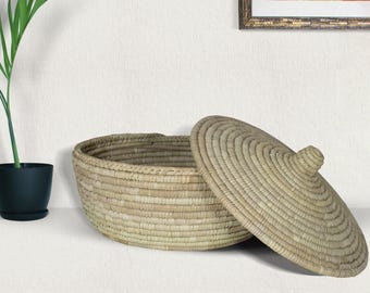 Puede incluir: Una cesta grande, redonda y tejida con tapa. La cesta está hecha de materiales naturales y tiene un color marrón claro. La tapa es ligeramente abovedada y tiene un pequeño pomo en la parte superior.