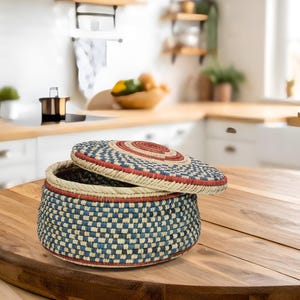 May include: A round, lidded storage basket featuring a woven pattern in shades of blue, cream, and red. The basket has a flat base and a matching circular lid that is slightly tilted, revealing the dark interior of the container.
