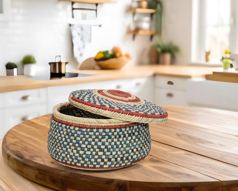 May include: A round, lidded storage basket featuring a woven pattern in shades of blue, cream, and red. The basket has a flat base and a matching circular lid that is slightly tilted, revealing the dark interior of the container.