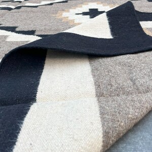 May include: A folded, patterned blanket in shades of gray, black, and cream. The design features geometric shapes and a black border. The blanket's texture is visible, suggesting a woven or knitted material. The overall aesthetic is rustic and cozy.